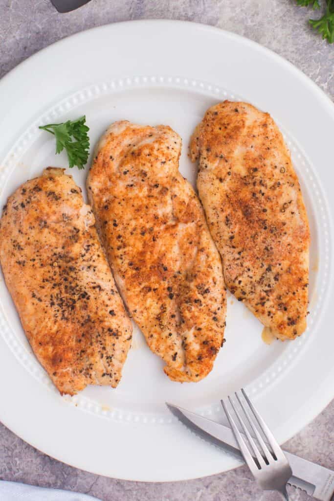 Three air fryer grilled chicken breasts on a white plate with a fork and knife toward the bottom.