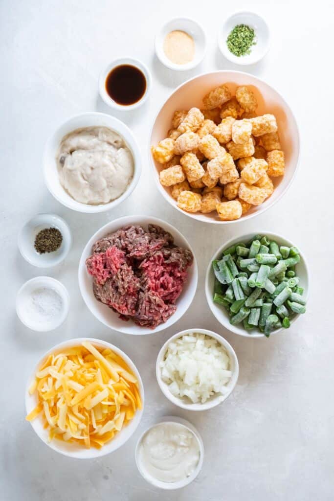 Ingredients needed to make green bean tater tot casserole: ground beef, onion, garlic powder, parsley, salt, pepper, worcestershire sauce, cream of mushroom soup, sour cream, frozen green beans, cheddar cheese, and tater tots.