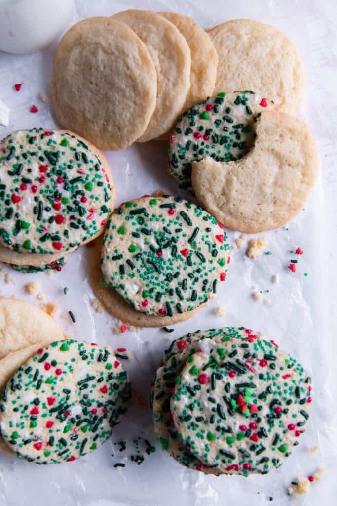 Sugar cookies without baking soda on a counter with some with Christmas sprinkles baked into them and one with a bite taken out of it.