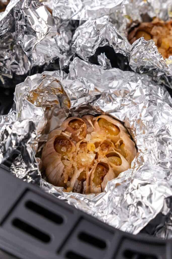 Close up of the roasted garlic in the air fryer basket with the foil partially unwrapped so you can see it fully roasted.