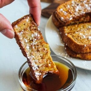 air fryer french toast sticks being dipped into a small bowl of maple syrup.
