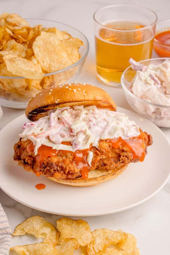 Buffalo chicken sandwich recipe with coleslaw mixture on top with the bun halfway off on a white plate.