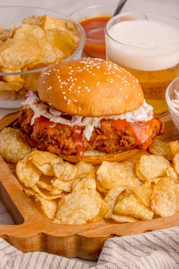 Buffalo chicken sandwich on a cutting/serving board with potato chips.