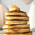 Pouring maple syrup over a stack of sweet cream pancakes.