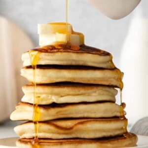 Pouring maple syrup over a stack of sweet cream pancakes.