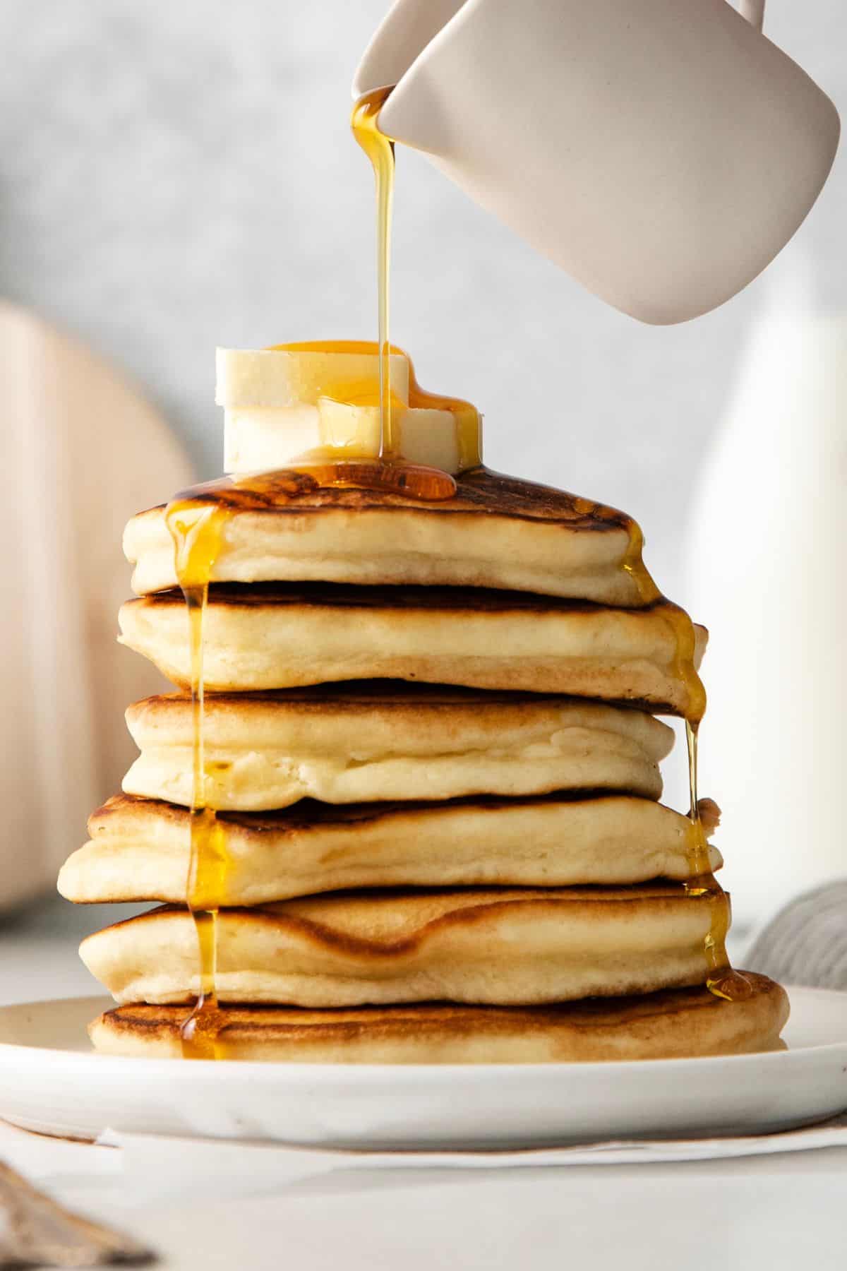 Pouring maple syrup over a stack of sweet cream pancakes.