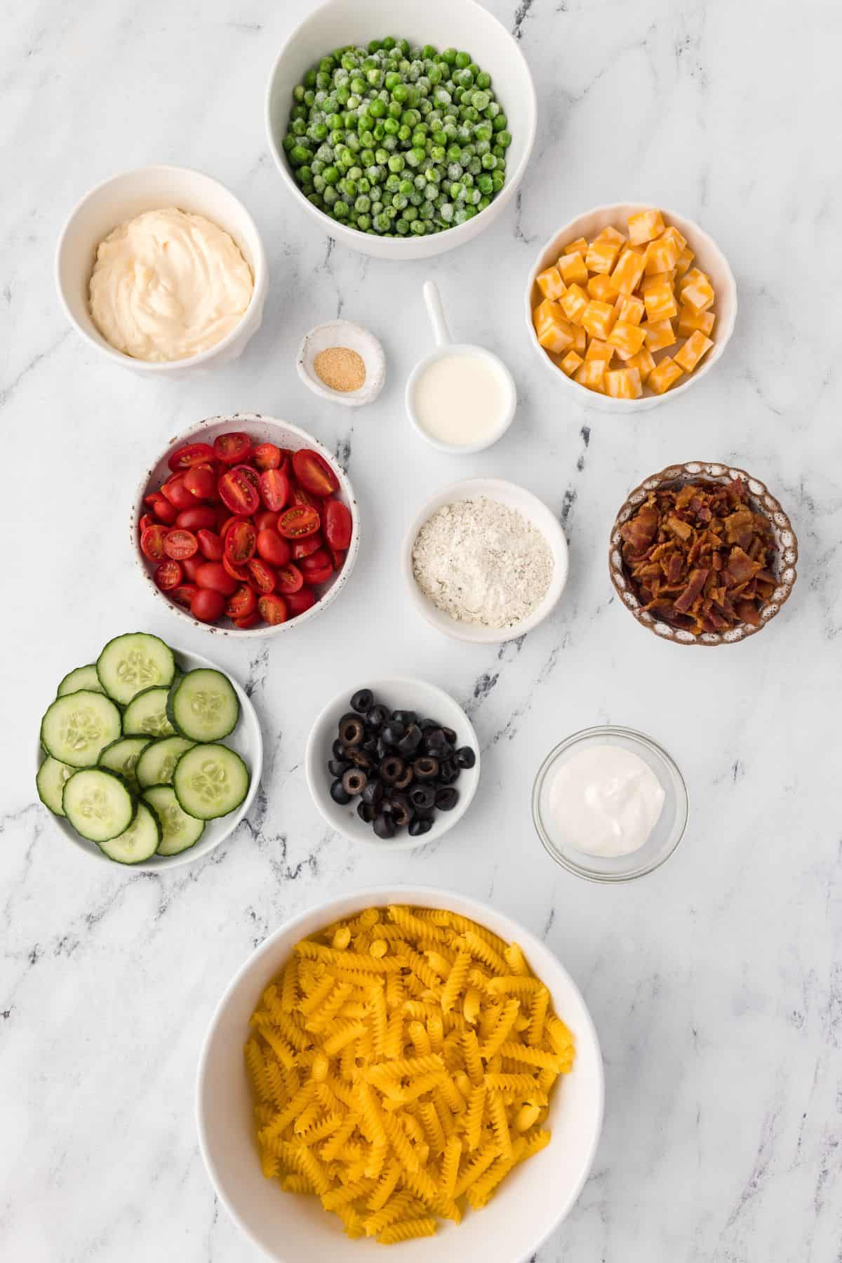 Ingredients needed to make bacon ranch pasta salad: bacon, rotini pasta, mayo, ranch seasoning, garlic powder, heavy whipping cream, sour cream, peas, olives, cherry tomatoes, colby monterey jack cheese, and cucumber.