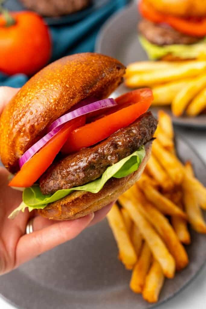 Closeup view of a fully dressed hamburger that has been prepared in a Ninja Foodi with a gray plate holding french fries in the background.