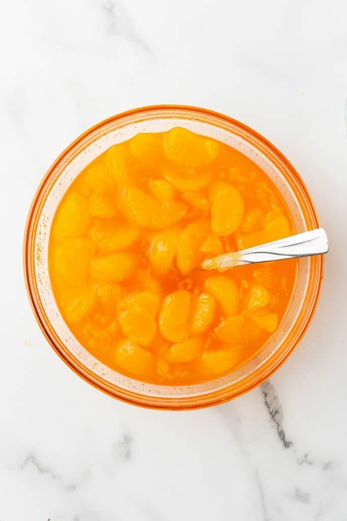 Overhead view of orange jello with mandarin oranges in a clear bowl with a spoon.