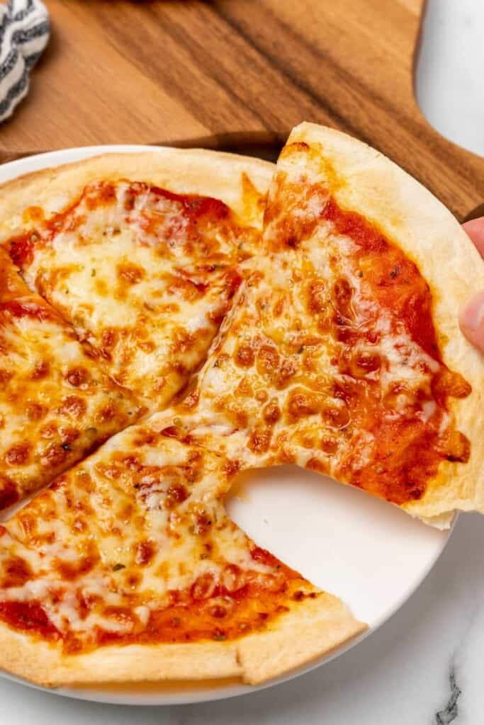 Lifting a slice of air fryer tortilla pizza off a white plate.