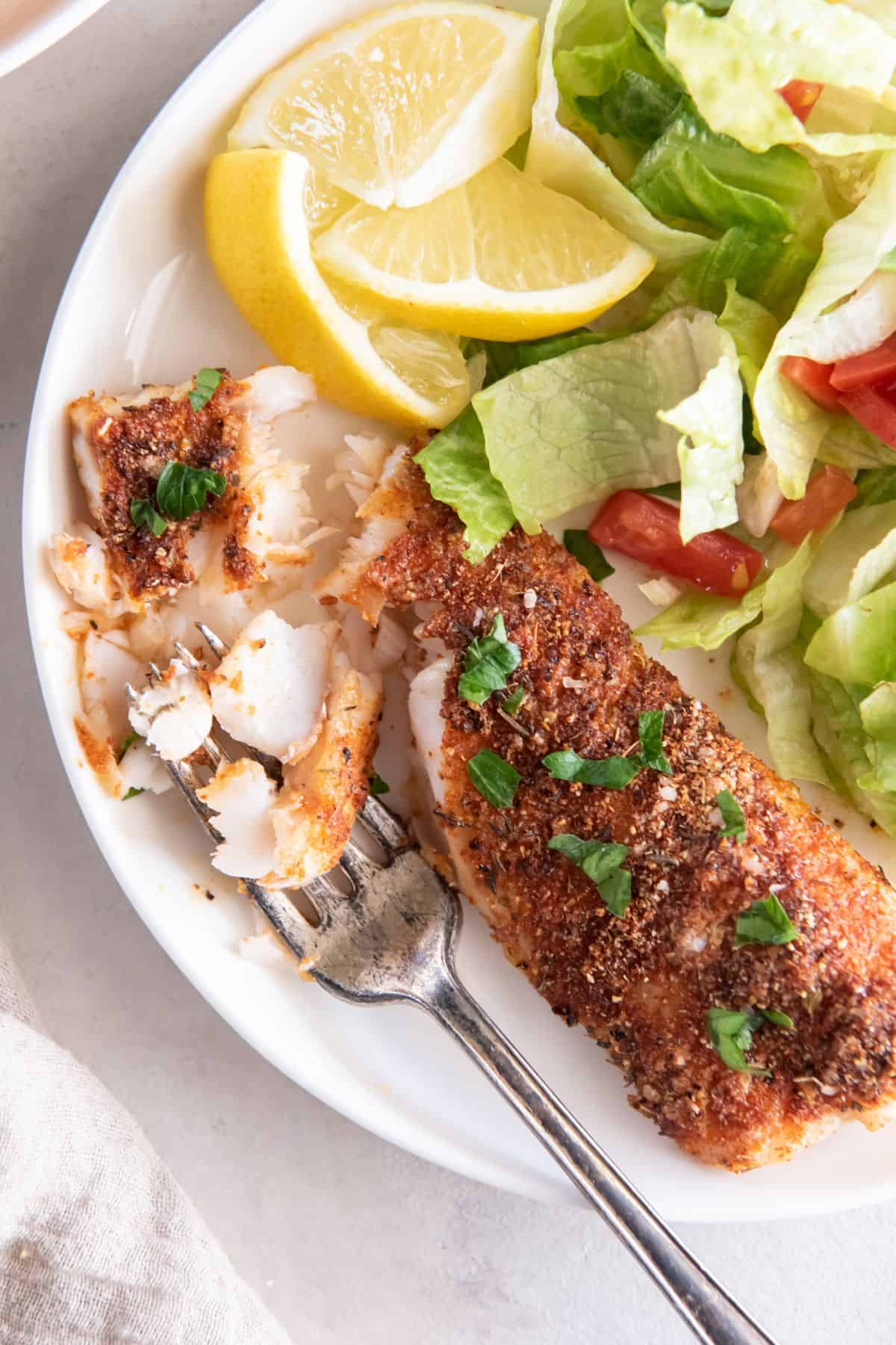 Seasoned cod on a plate with pieces taken off with a fork to see the flakiness inside the fish.
