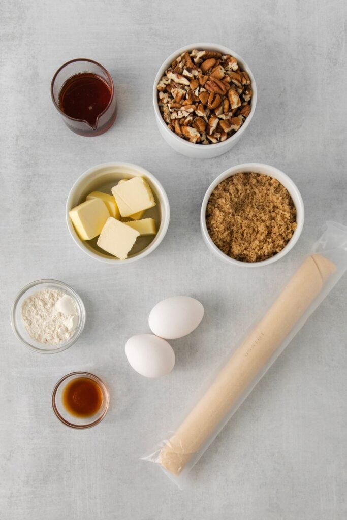 Ingredients needed to make a pecan pie without corn syrup: pie crust, brown sugar, butter, maple syrup, eggs, flour, vanilla extract, and pecans.