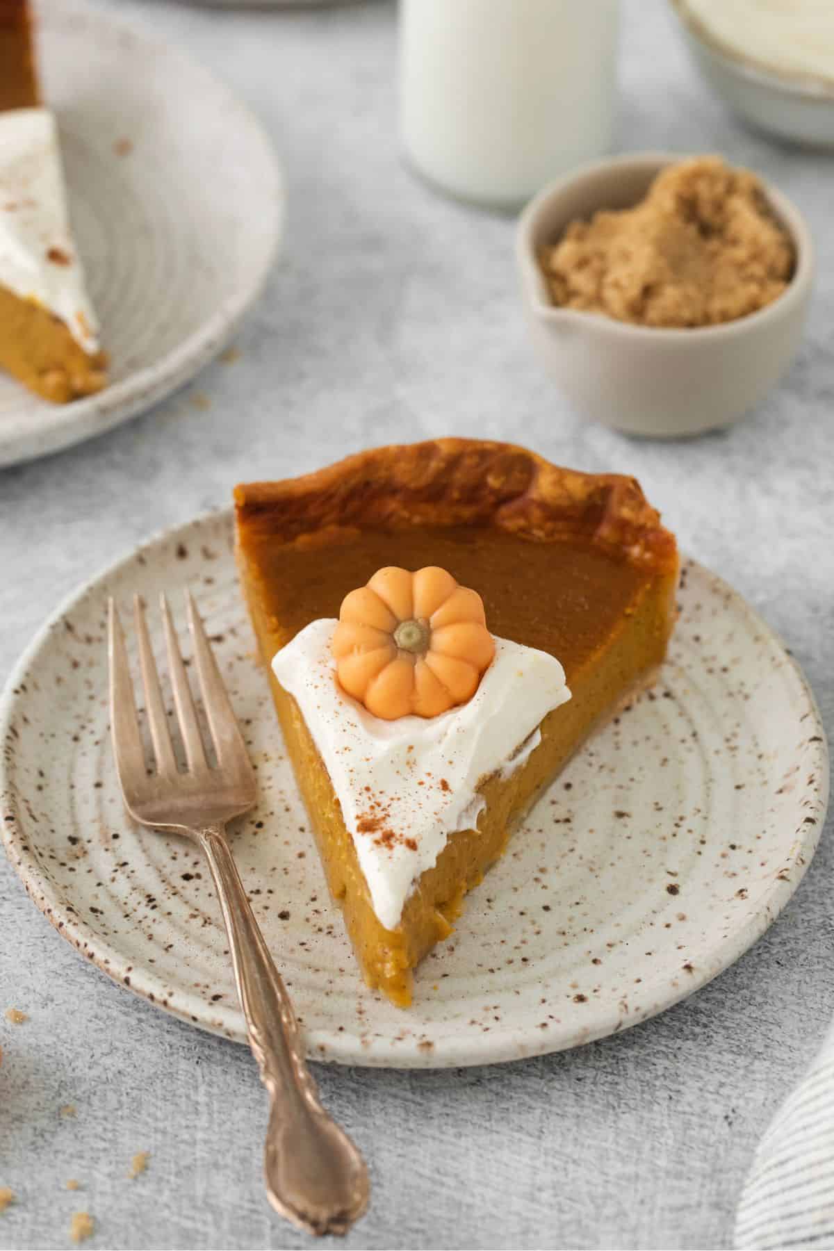 Slice of pumpkin pie with whipped cream and decorative pumpkin on top on a white plate with a fork.