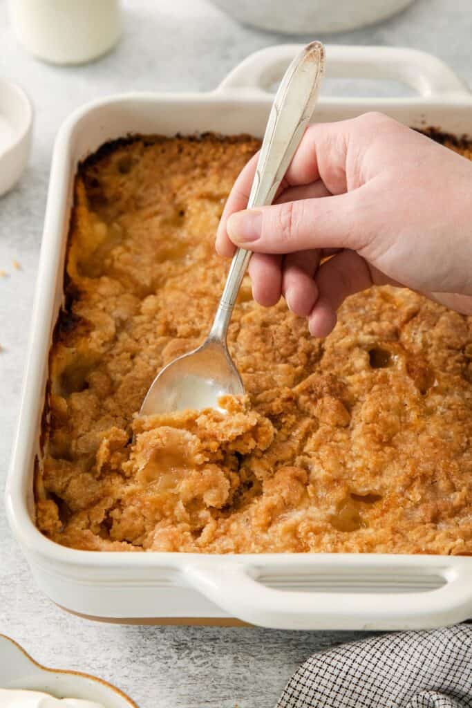 Close up of baking dish with baked 4 ingredient peach cobbler with a spoon about to take out a piece.