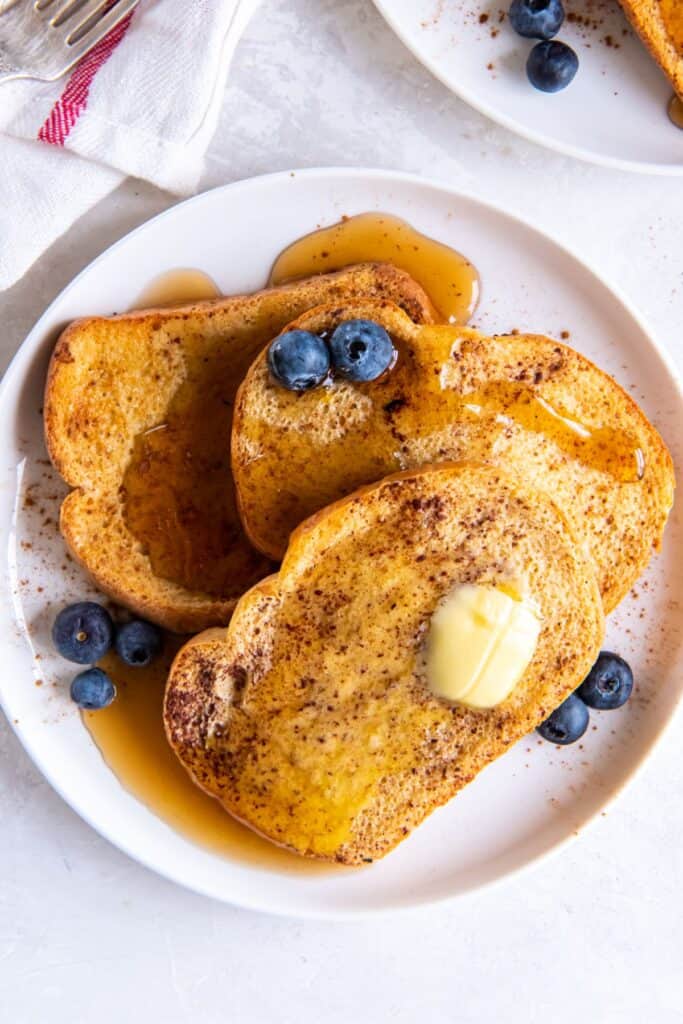 Overhead shot of air fryer french toast on a white plate with butter, maple syrup, and blueberries on them.