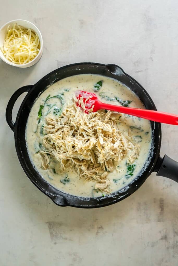 Shredded chicken mixed into alfredo sauce in a pan.