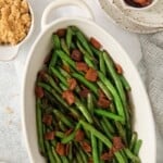 Vertical view of green beans with bacon in an oblong serving dish on a white tablecloth.