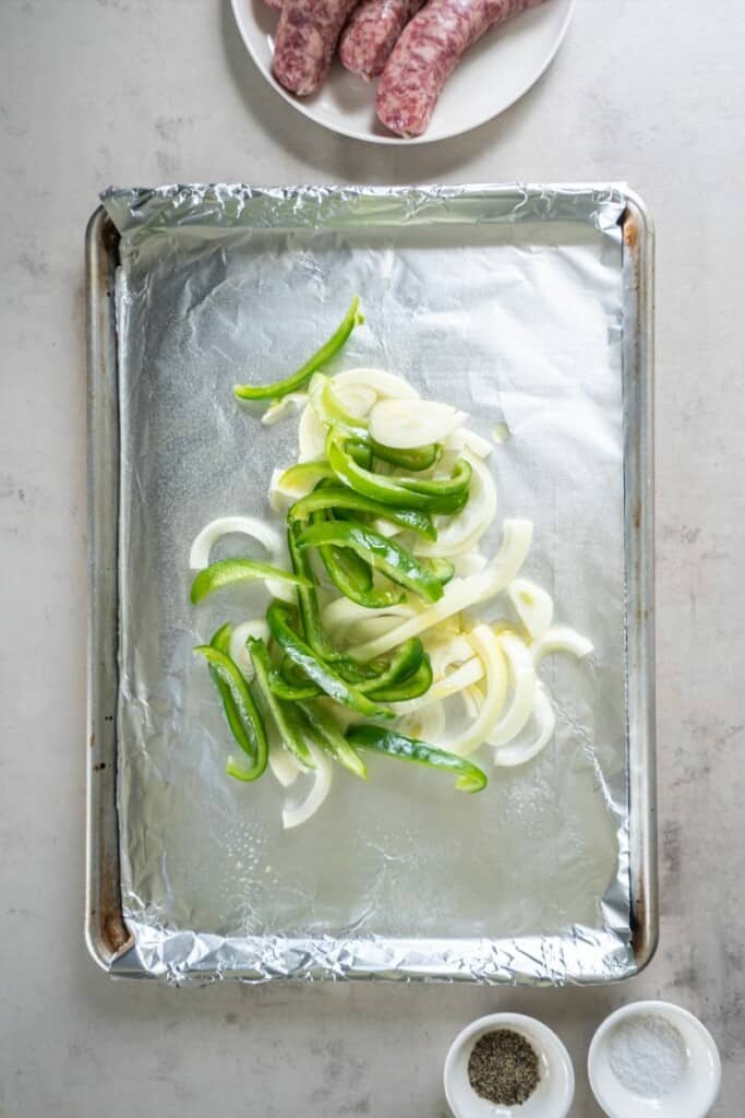 Onions and peppers on a foil lined baking sheet with brats and salt & pepper in the background.