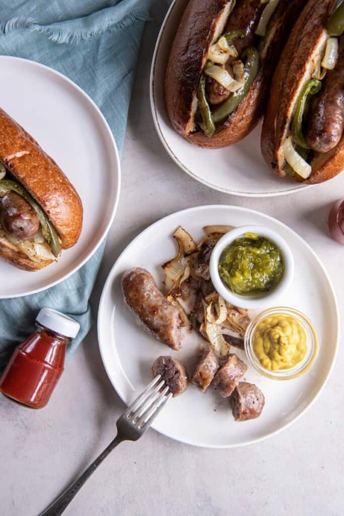 Overhead view of a sliced brat on a plate with mustard & relish with additional brats in buns in the background.