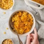 A bowl of macaroni corn casserole surrounded by cheese and the casserole dish.