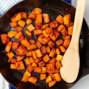 Diced butternut squash that has been sauteed in a skillet with a wooden spoon.