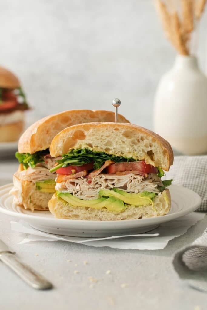 A turkey bacon avocado sandwich sliced in half on a white plate.