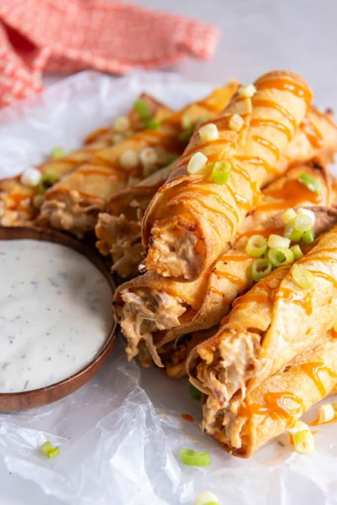 A stack of prepared buffalo chicken taquitos with green onions on top and dipping sauce next to the stack,