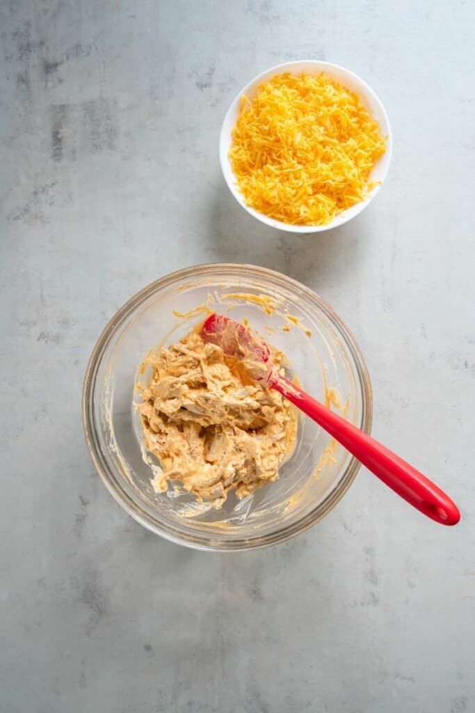 Combining shredded chicken with buffalo sauce in a bowl. A bowl of grated cheese next to it.