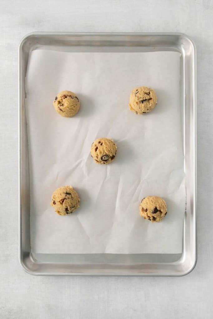 Five cookie dough balls on a baking sheet lined with parchment paper.