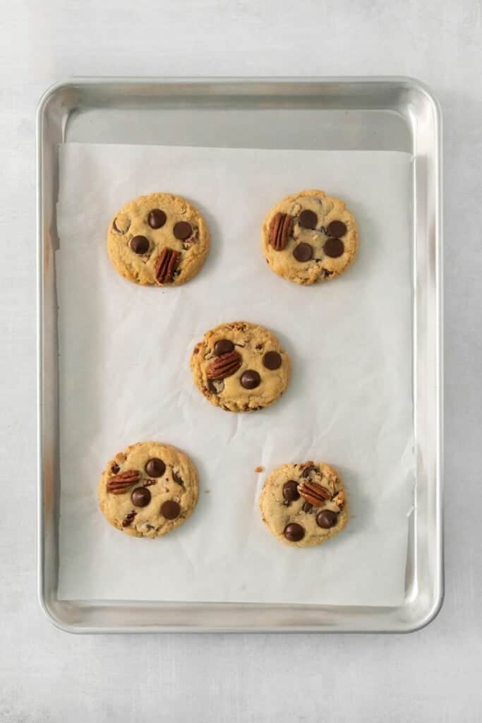 Five baked pecan chocolate chip cookies on a baking sheet lined with parchment paper.