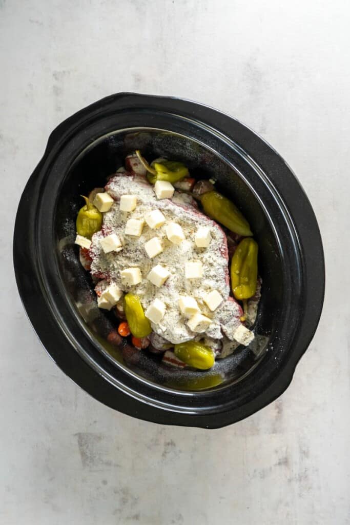 Seasonings, peppers and butter added to pot roast and vegetables in a black slow cooker.
