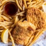 A basket of prepared fish and chips with lemon wedges and dipping sauce.