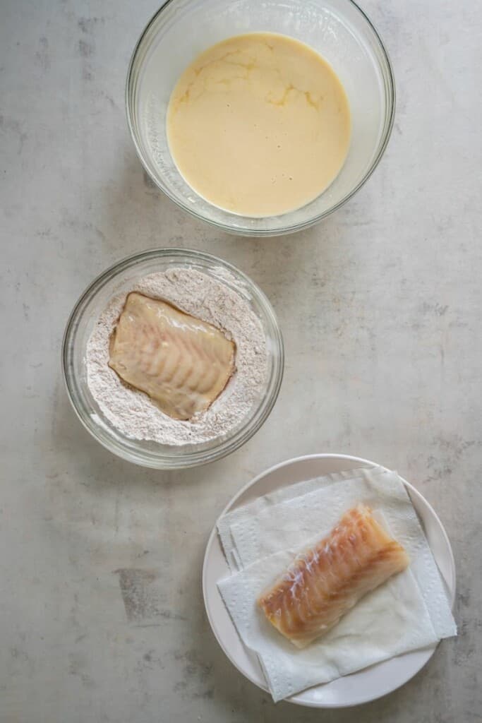 A bowl of fish batter, a bowl of flour with one cod filet, a plate with a second cod filet.