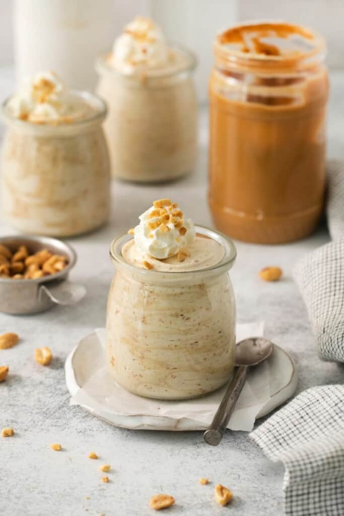 A few jars of peanut butter mousse topped with whipped cream. A jar of peanut butter in the background.