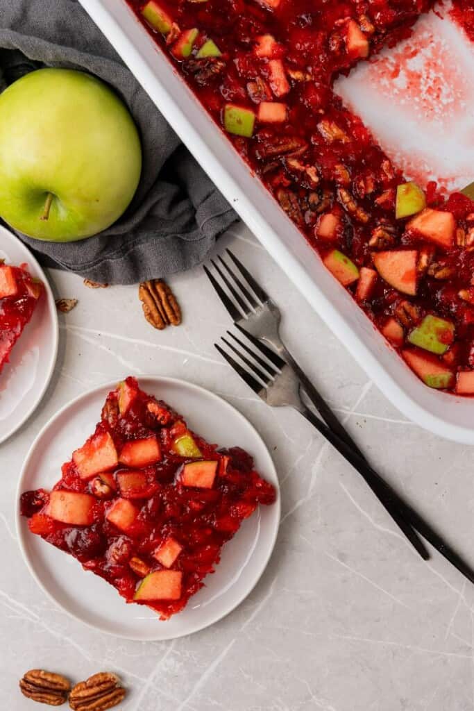 A slice of cranberry jello salad on a saucer next to the dish holding the remaining salad.