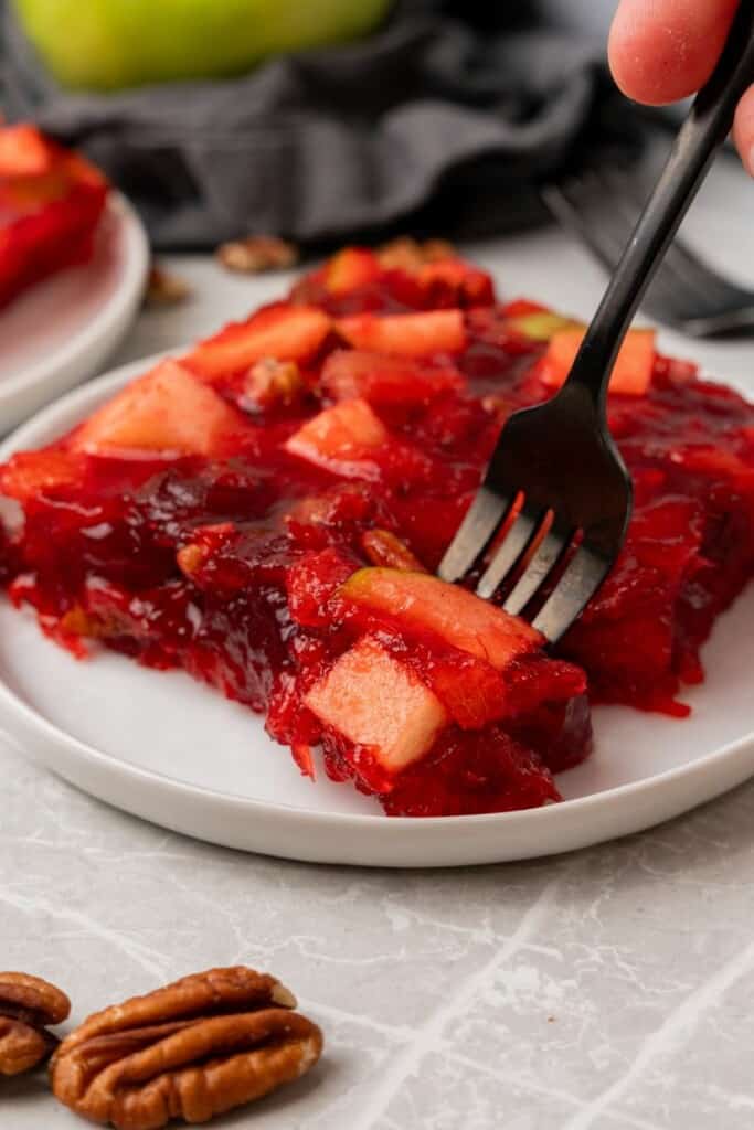A fork taking a bite of jello salad from the serving on a white saucer.