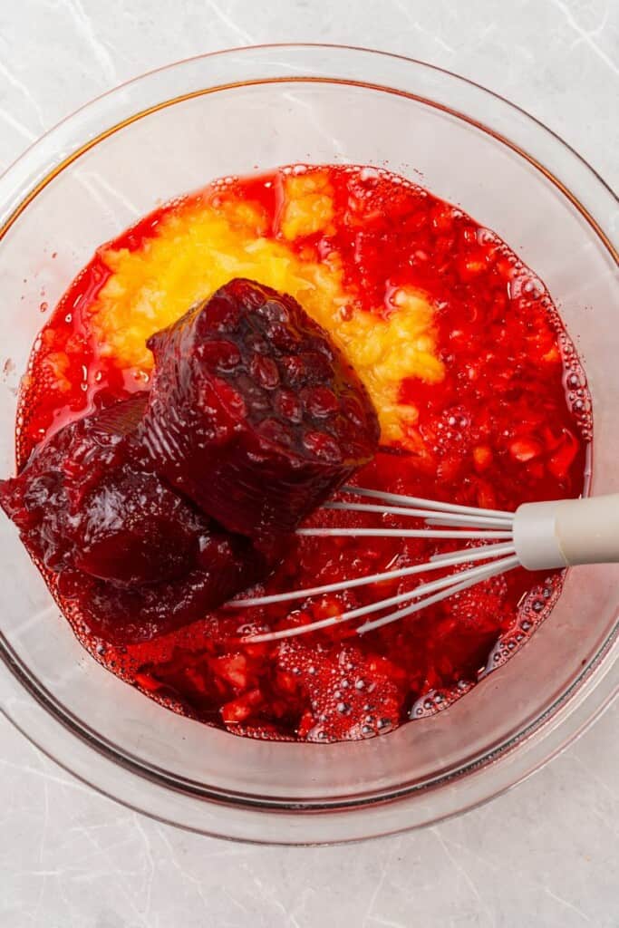 Ingredients for cranberry jello salad in a clear mixing bowl with a whisk ready to be combined.