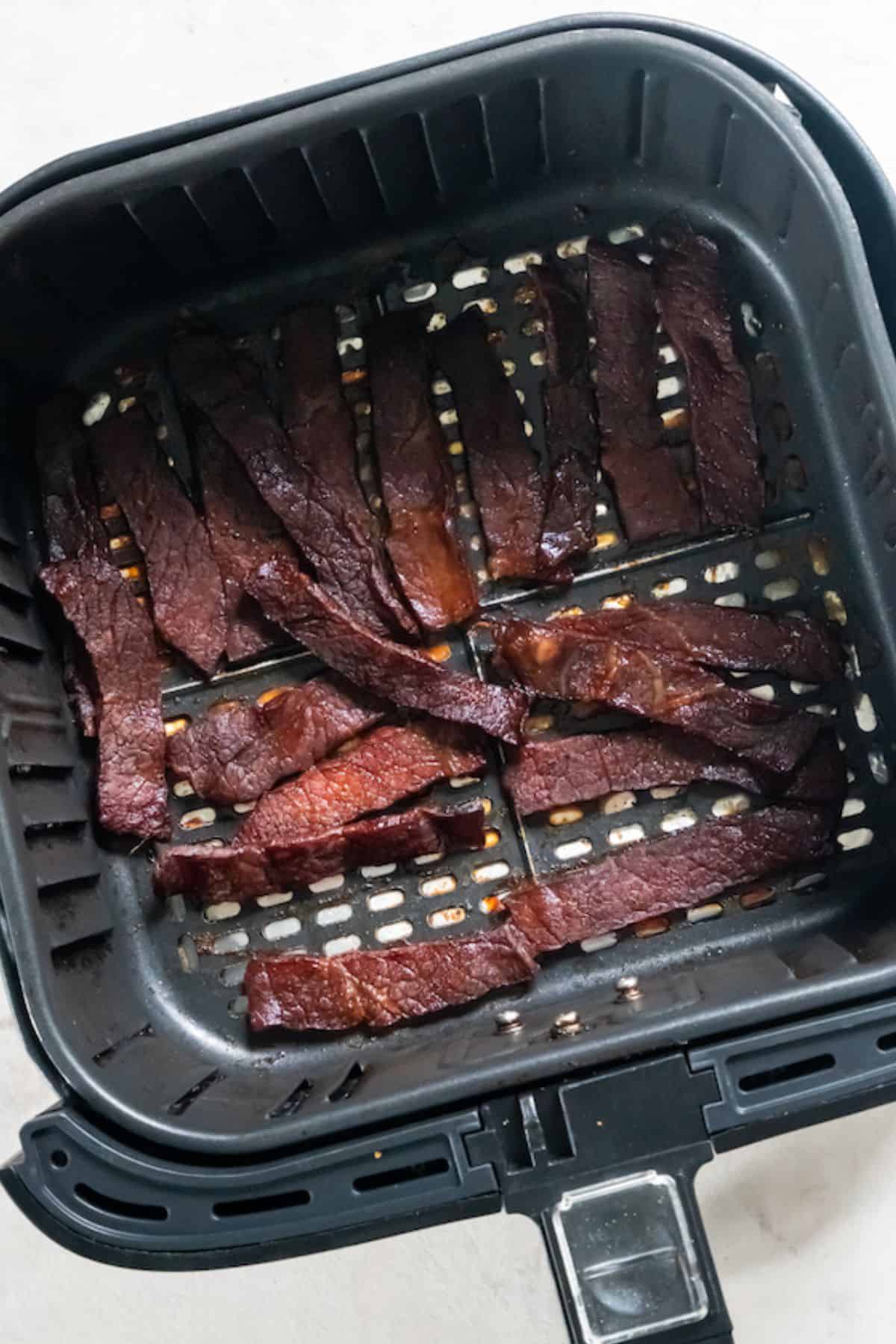 A closeup view of beef jerky slices in a black air fryer basket.