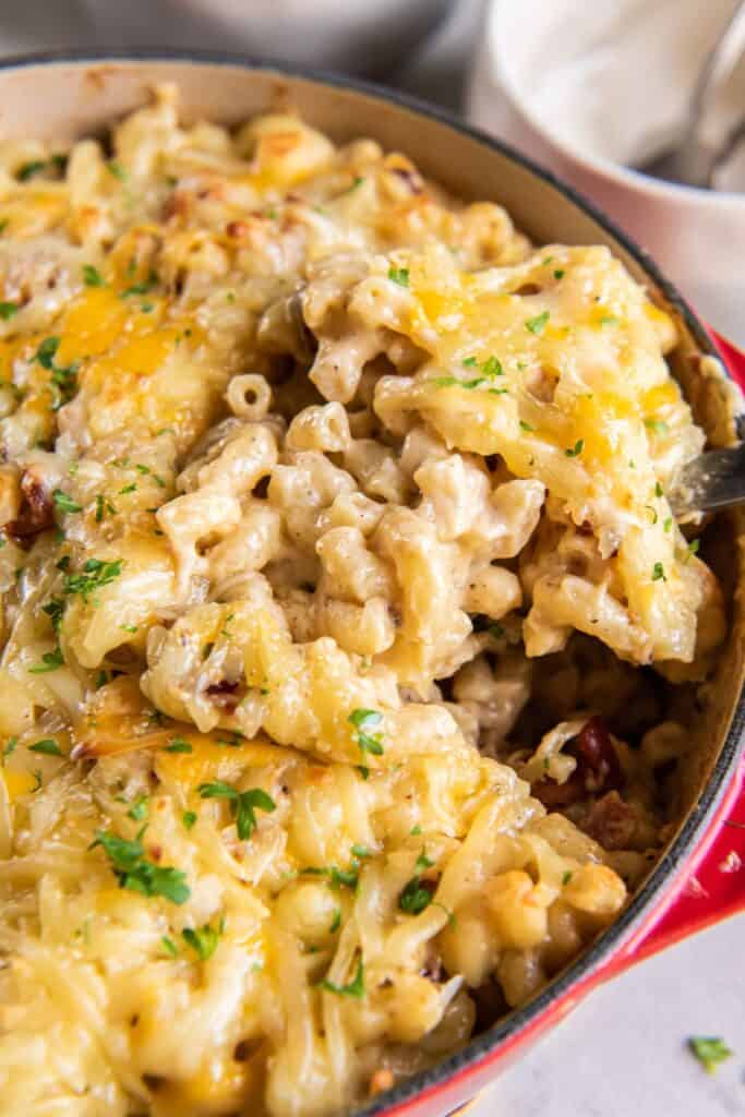 Dutch oven mac and cheese in a dutch oven pot being scooped up by a serving spoon so you can see the cheesiness and bacon.