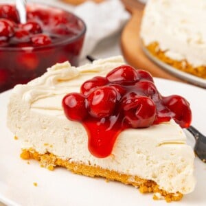 A slice of no bake cheesecake with Cool Whip on a white plate with cherry pie filling on top spilling off the sides.