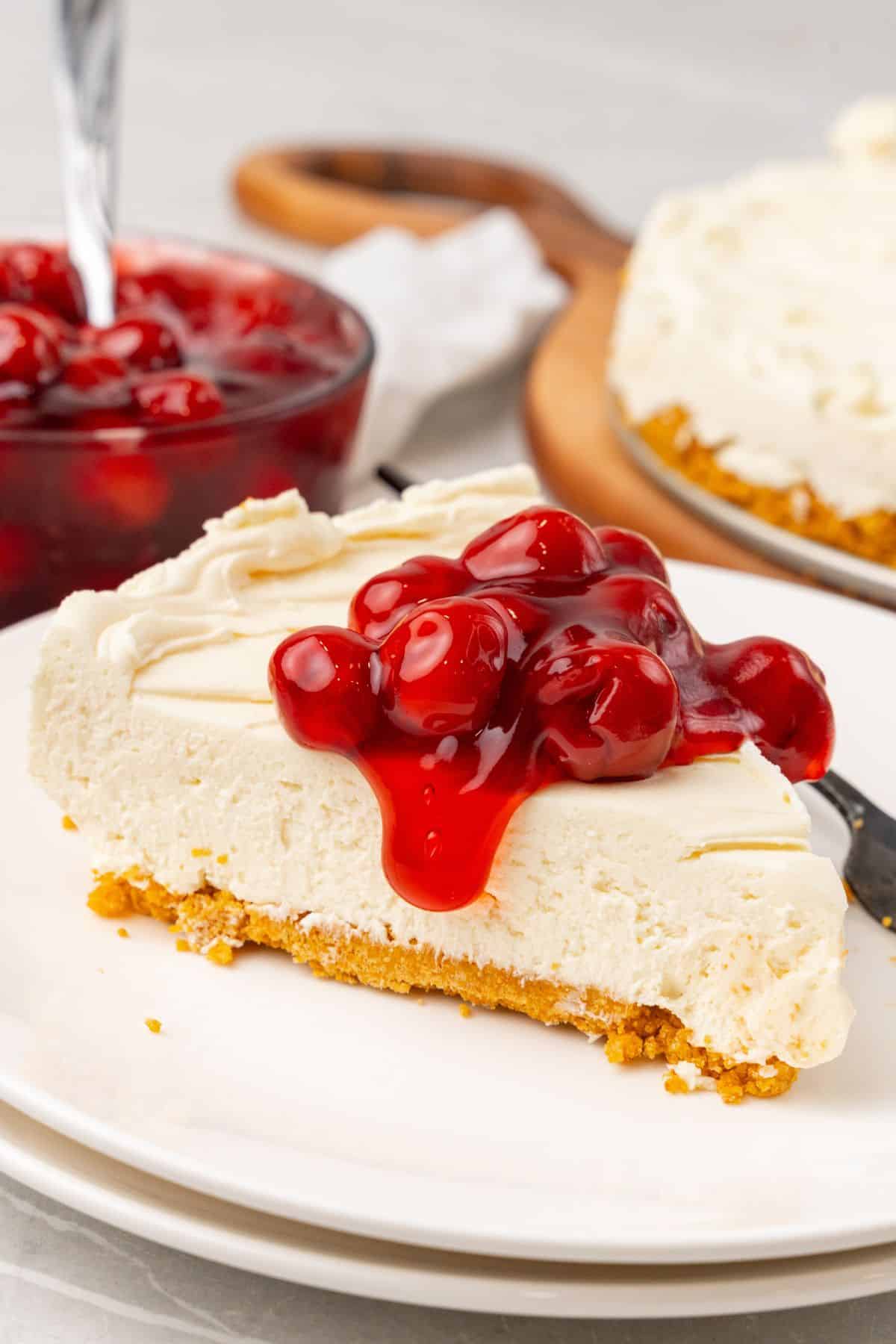 A slice of no bake cheesecake with Cool Whip on a white plate with cherry pie filling on top spilling off the sides.