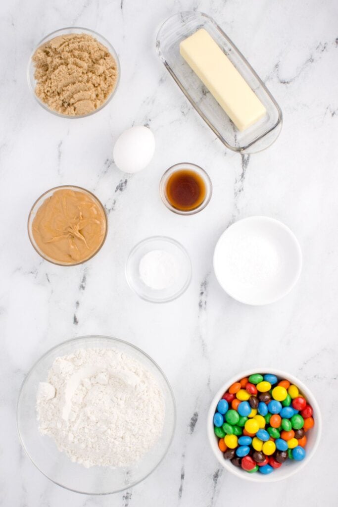 Ingredients needed to make peanut butter M&M cookies: butter, brown sugar, egg, creamy peanut butter, vanilla extract, baking soda, flour, peanut butter M&Ms, and salt.