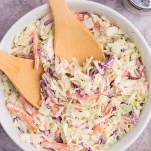 Easy coleslaw recipe in a white bowl with wooden utensils for serving.
