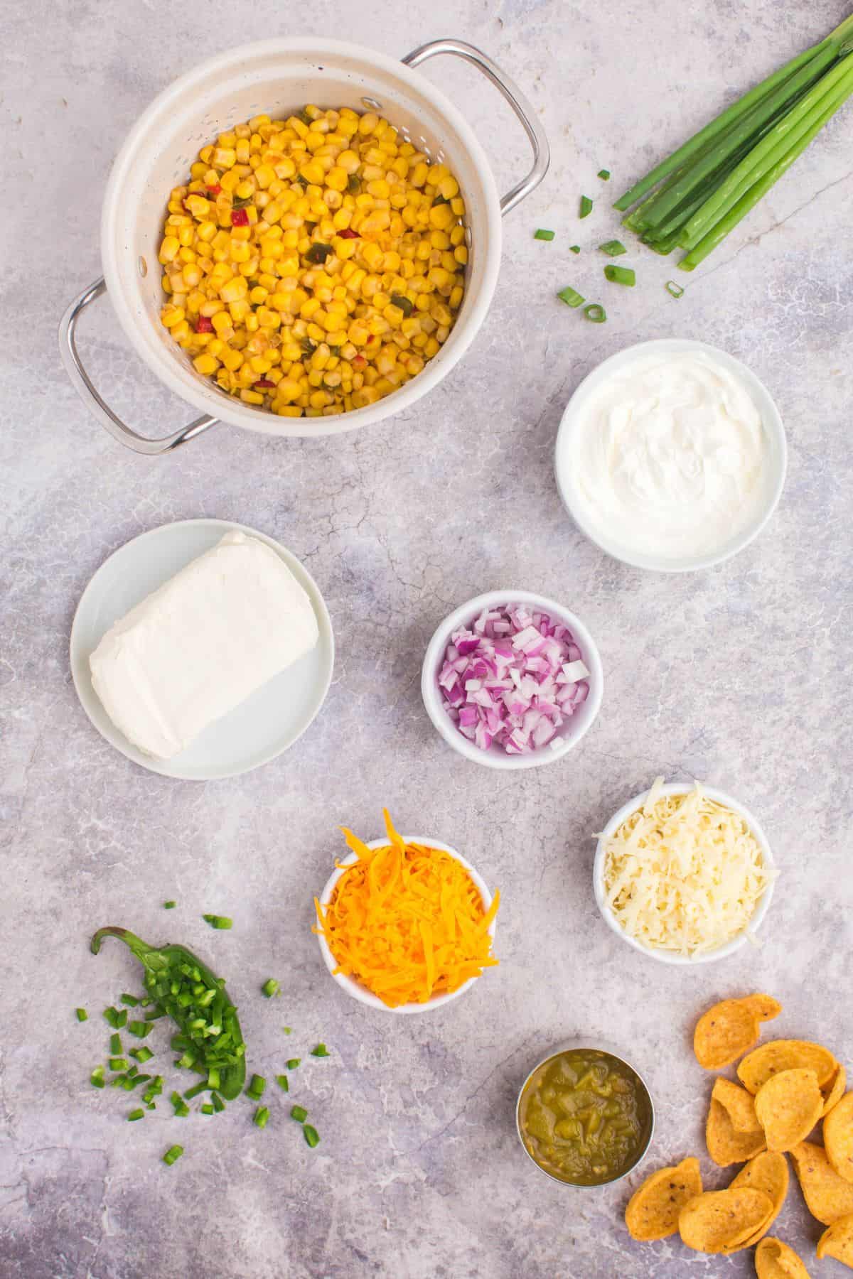 Ingredients needed to make crack corn dip: fiesta corn, cream cheese, sour cream, pepper jack cheese, cheddar cheese, green chiles, jalapeno pepper, red onion, green onions, and fritos.