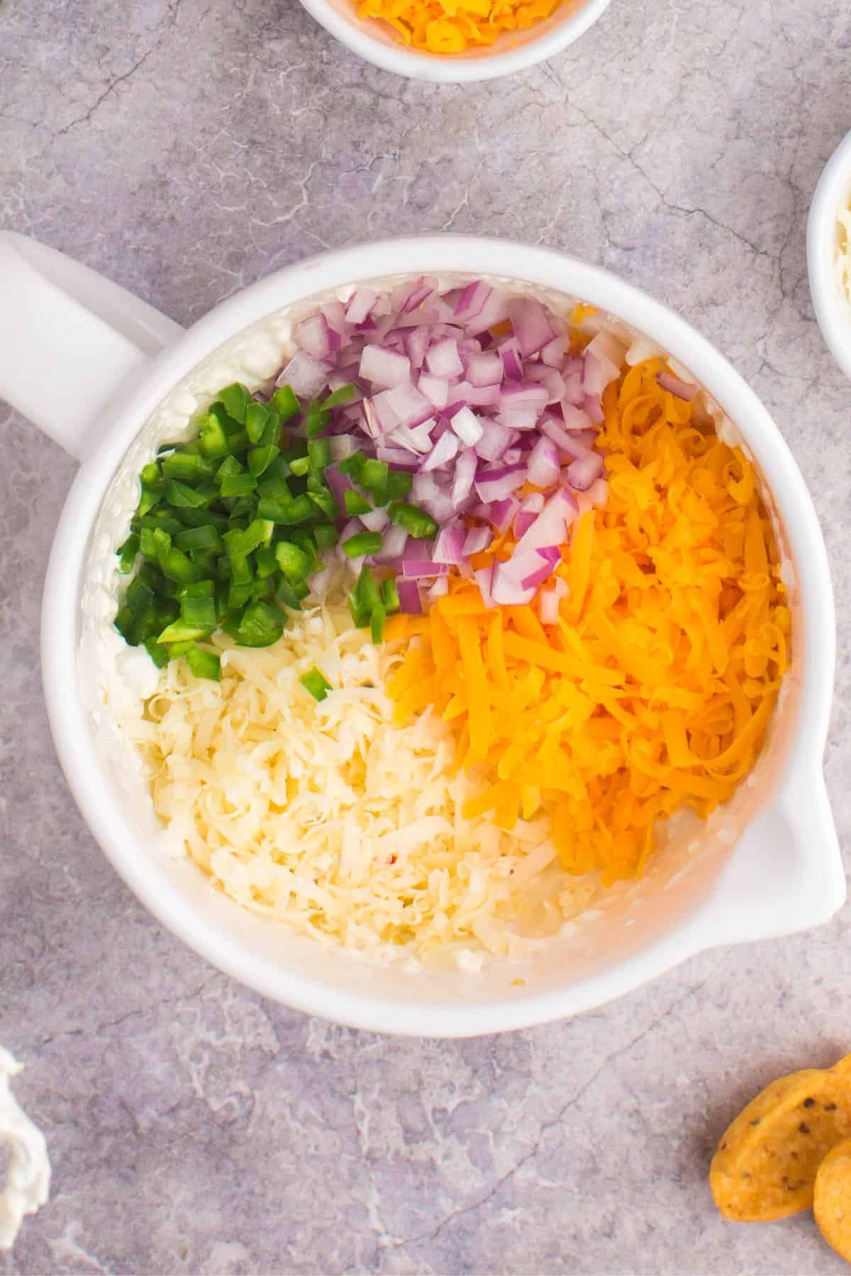 The cheese, pepper, onion, and green onion being added to the mixing bowl to make the corn dip with fritos.