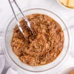 Pulled pork in a bowl with prepped bowls of BBQ sauce, potato chips and hamburger buns surrounding it