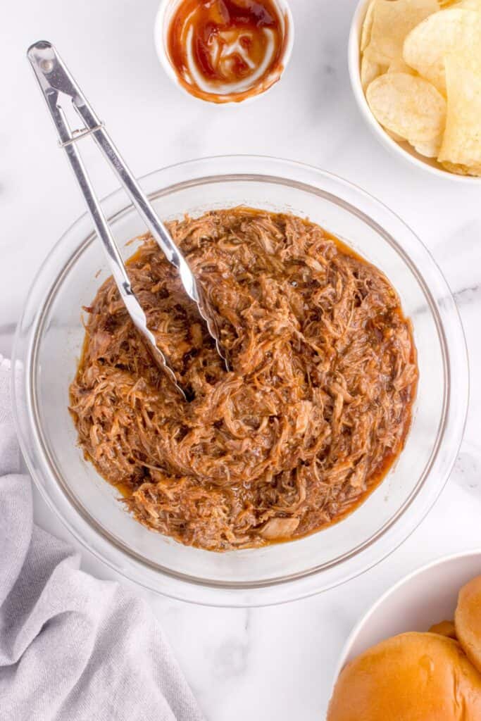 Pulled pork in a bowl with prepped bowls of BBQ sauce, potato chips and hamburger buns surrounding it