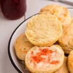 Serving bowl with biscuits piled up. Top biscuit is halved and spread with butter and strawberry jam.