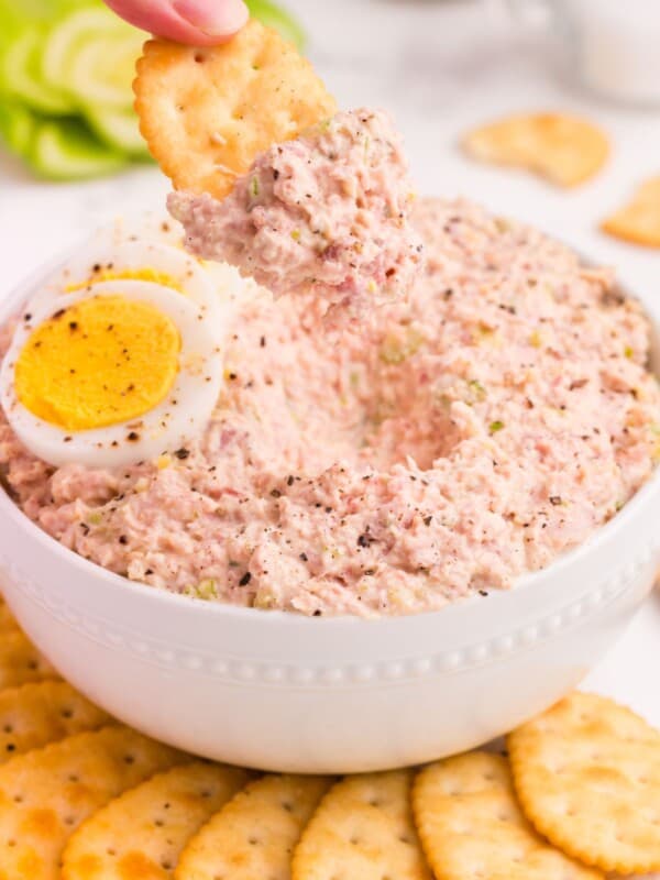 Ham salad in a bowl surrounded by crackers. A hand is scooping up ham salad with a cracker.