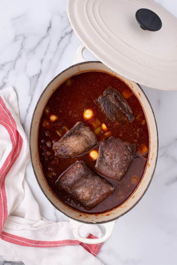 Dutch oven with beef short ribs. The lid is balanced on the edge of the pot.